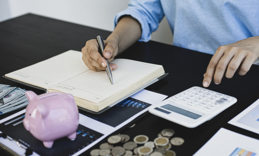 Person calculating expenses and writing notes beside coins, cash, and a piggy bank
